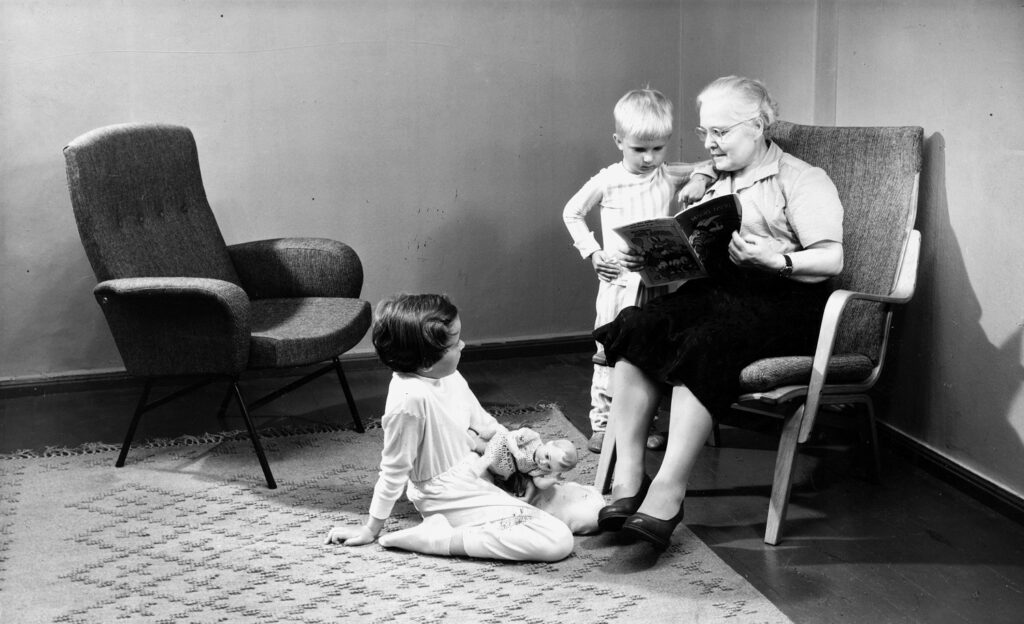 Vanhus istuu olohuoneessa Askon tuolissa sylissään lapsi. Toinen lapsi istuu lattialla ja katsoo heihin.