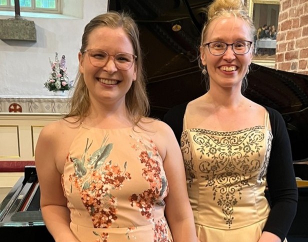 Kaksi muusikkoa hymyilee flyygelin edessä ja katsoo kmeraan. He ovat pukeutuneet vaaleisiin esiintymismekkoihin.